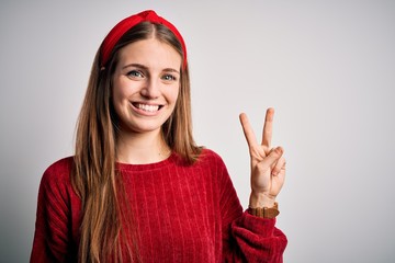 Young beautiful redhead woman wearing red casual sweater and diadem over yellow background showing and pointing up with fingers number two while smiling confident and happy.