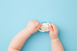 Infant hands holding white soother on light blue floor background. Pastel color. Closeup. First baby toy. Top down view.