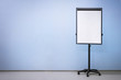 Flip chart board on wheels with a blank sheet of paper on a light blue wall background, copy space. Empty room for educational activities.