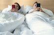 © Space_Cat - Young married couple using their mobile phone in bed ignoring each other as strangers. Relationship and communication problems and internet social network addiction