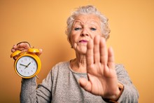 Senior Beautiful Woman Holding Alarm Clock Standing Over Isolated Yellow Background With Open Hand Doing Stop Sign With Serious And Confident Expression, Defense Gesture