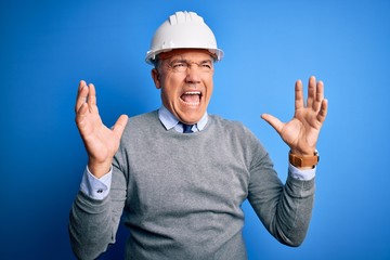 Poster - middle age handsome grey-haired engineer man wearing safety helmet over blue background crazy and ma