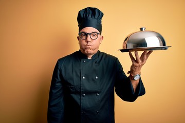 Poster - middle age handsome grey-haired waiter man wearing cooker uniform and hat holding tray puffing cheek