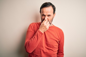 Wall Mural - Middle age hoary man wearing casual orange sweater standing over isolated white background smelling something stinky and disgusting, intolerable smell, holding breath with fingers on nose. Bad smell