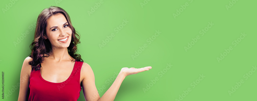 Happy smiling woman giving, holding or showing something or product or copy space for some text or slogan, green background. Brunette model in casual red clothing posing at studio. - obrazy, fototapety, plakaty 