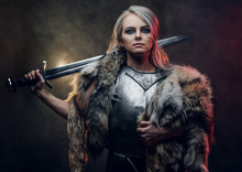 Portrait Of A Beautiful Warrior Woman Holding A Sword Wearing Steel Cuirass And Fur. Fantasy Fashion. Studio Photography On A Dark Background. Cosplayer As Ciri From The Witcher.