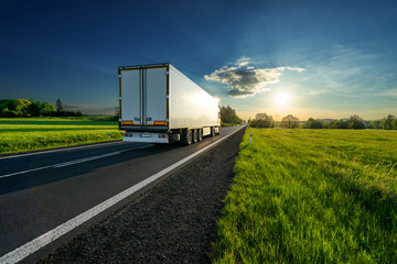Wall Mural - white truck driving on the asphalt road in a spring rural landscape at sunset