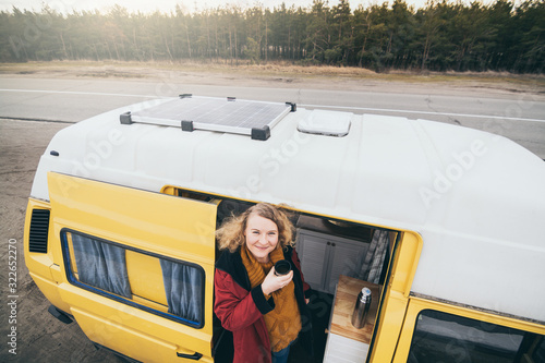 Young blond woman looking out of camper van with solar panel on the roof top and pine forest on the background
