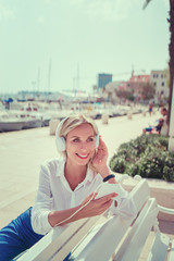 Wall Mural - Enjoying the sound. Happy young woman with earphones listening music on smartphone while sitting on city embankment sea promenade.