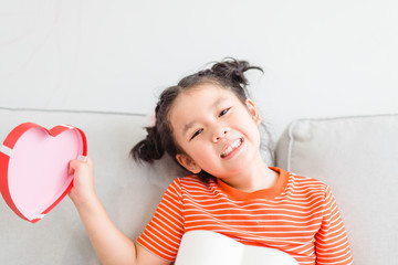 Wall Mural - Little asian girl Happy smiley excited face and open red gift box in heart shape in sofa at home on white wall background.Child girl holding gift box in Valentine day, Christmas and New year concept.