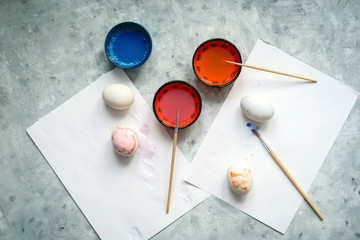 Wall Mural - top view selective focus cups with dye, brushes and white eggs on a concrete background, painting eggs for Easter diy concept