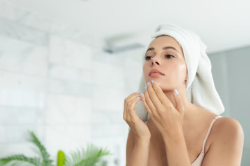 Young beautiful woman wearing bath towel and underwear standing in bathroom and looking for or squeezing acne on chin. Mirror reflection. Ugly problem teen girl skin, pimples. Skincare and beauty