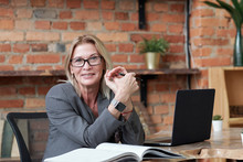 Portrait Of Smiling Mature Woman In Glasses Sitting At Desk With Laptop In Loft Office And Examining Notes In Accounting Book