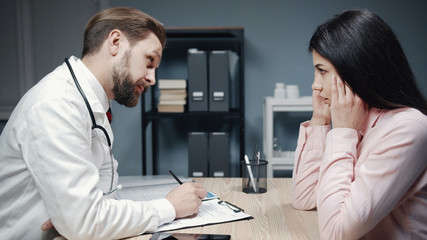 Mature male doctor works with female patient, listening complaints and making medical records