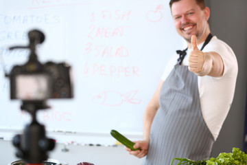 Cheerful Blogger Chef Showing Thumbup Gesture Joke. Cook in Apron Joking with Green Cucumber in Hand. Man Recording Culinary Healthy Recipe for Blog. Ripe Organic Cuke. Blackboard with Ingredient