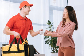 Wall Mural - Smiling delivery man with thermo bag giving food container to attractive girl at home