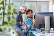 © Sanja - Two co-workers talking on a work break at the office. The man is showing something to the woman on his phone while she sits leaning against the window sill.
