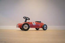 Vintage Red Toy Racing Car Seen From Side, Against Grey Wall And On Wooden Floor