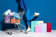 © LIGHTFIELD STUDIOS - cropped view of african american woman with shopping cart full of gifts near shopping bags on blue background