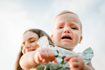 Portrait of a little beautiful baby girl crying and mom on nature on summer day. The playing in the park with parents. Close Up. The concept of family holiday and time together.