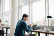 © Maskot - Young businessman using smart phone while sitting with laptop at desk in office