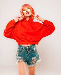 Young beautiful hipster bad girl in trendy red summer hoodie and earring in her nose.Sexy carefree woman in pink wig posing in studio on gray background.Hot model in sunglasses