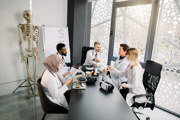 Multi ethnic group of medical students or young reseachers having a discussion during the conference meeting with documents, microscope and vr goggles at the table in modern office or lab