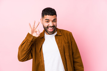 Wall Mural - Young latin man against a pink background isolated winks an eye and holds an okay gesture with hand.