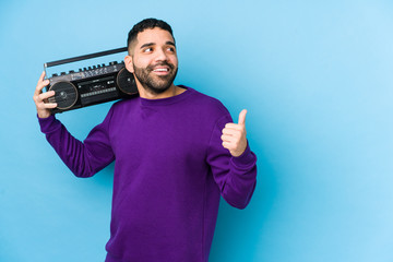 Wall Mural - Young arabian man holding a radio cassette isolated Young arabian man listening music points with thumb finger away, laughing and carefree.