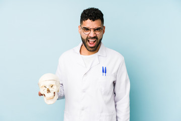 Wall Mural - Young doctor latin man holding a skull isolated screaming very angry and aggressive.