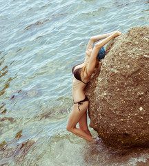 Wall Mural - Beautiful slim young woman in a black bikini swimsuit resting on a rocky beach