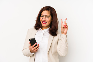 Middle age latin woman holding a mobile phone isolated joyful and carefree showing a peace symbol with fingers.