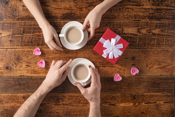 Valentine's Day celebration concept. A nice gift for your loved one. Hands of man and woman with coffee mugs on a wooden table background. Copy space. Flat lay. Close-up.