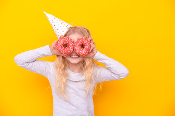 Crazy cheerful blonde girl in birthday hat smiling, having fun and looking through two red donuts on her eyes. Sweets. Yellow studio background.