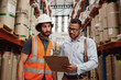 © StratfordProductions - Portrait of Warehouse owner talking to friendly young banker - warehouse manager wearing formal clothing talking to loader showing order list in aisle between tall shelves with packed goods