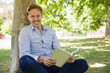 © Mangostar - Smiling young man leaning on tree and reading book. Handsome cheerful student studying in spring park. Education, leisure concept