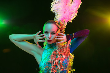 Graceful. Beautiful young woman in carnival, stylish masquerade costume with feathers on black background in neon light. Copyspace for ad. Holidays celebration, dancing, fashion. Festive time, party.