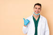 © luismolinero - Young dentist man holding tools isolated on beige background pointing to the side to present a product