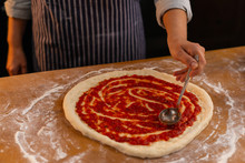 Female Chef Spreading With A Ladle Fresh Tomato Sauce Over Pizza Dough On A Wooden Table.