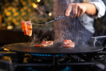 Wall Mural - Close up of a female chef flipping a sizzling, hot piece of bacon on a kitchen stove. 