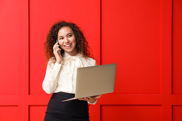 Young African-American businesswoman with laptop talking by phone on color background