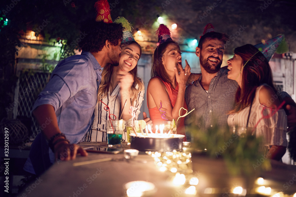 group of young friends celebrating a  birthday in the club with cake and candles at night.  birthday, party, fun, nighttime concept. - obrazy, fototapety, plakaty 