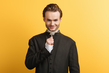 Wall Mural - young catholic priest holding cross on his necklace while smiling isolated on yellow