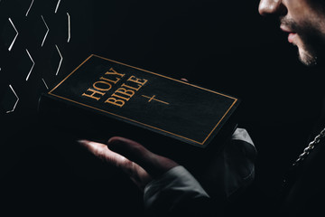 Poster - Cropped view of catholic priest holding holy bible near confessional grille in dark with rays of light