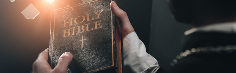 Poster - partial view of catholic priest holding holy bible near confessional grille in dark with rays of light, panoramic shot
