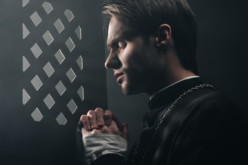 Wall Mural - young thoughtful catholic priest praying with closed eyes in dark near confessional grille with rays of light