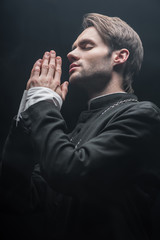 Sticker - low angle veiw of young concentrated catholic priest praying with closed eyes isolated on black