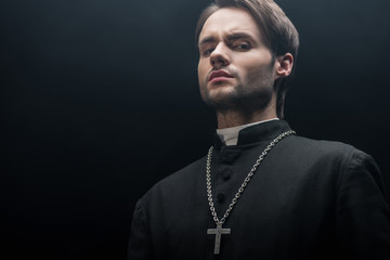 Wall Mural - low angle view of young tense catholic priest looking at camera isolated on black