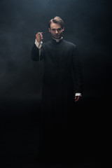 Sticker - full length view of confident catholic priest holding necklace with cross on black background with smoke