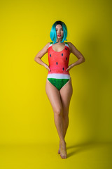 Wall Mural - Portrait of a woman in a swimsuit with a picture of a watermelon. Stylish girl in a colored short wig posing in the studio on a yellow background.